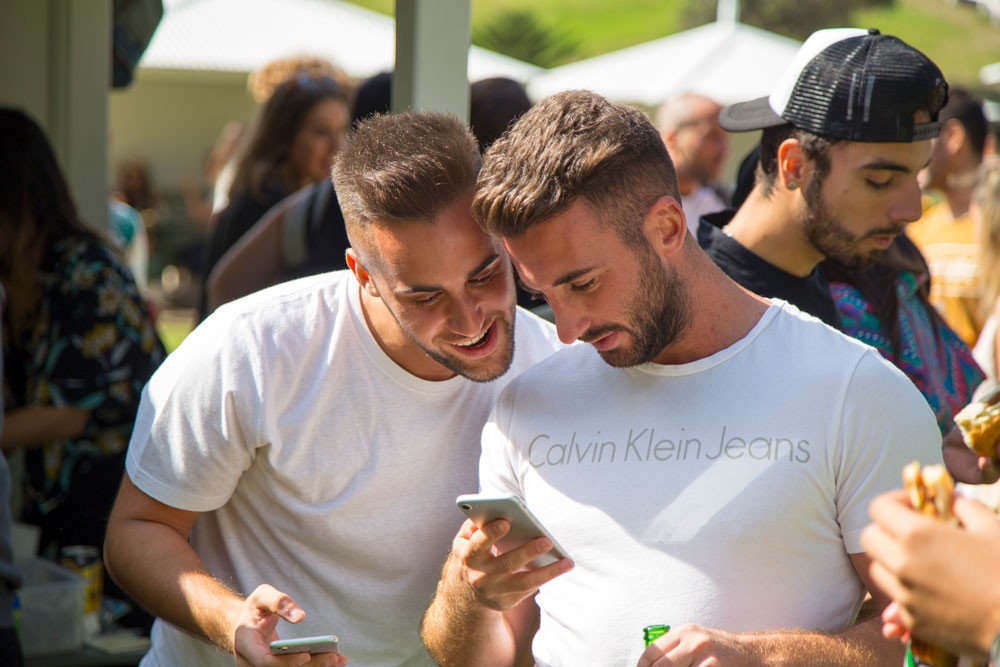 dos chicos mirando el teléfono de uno. Los dos llevan una camiseta blanca de manga corta y sonríen mientras miran la pantalla